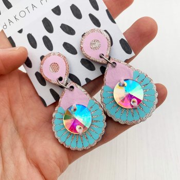 A pair of pastel pink and blue mini metallic earrings with iridescent gems are mounted on a black and white patterned, dakota rae dust branded card and held close to the camera lense by a white woman's hand