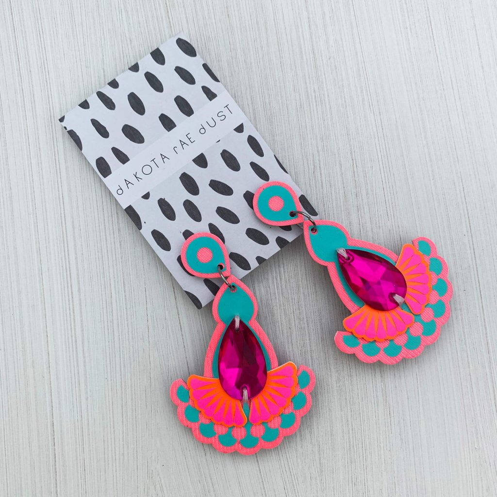 A pair of colourful hot pink and turquoise teardrop jewel earrings mounted on a black and white patterned, dakota rae dust branded card and seen against an off white background