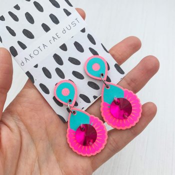 A pair of hot pink and turquoise mini jewel earrings mounted on a black and white patterned, dakota rae dust branded card are held close to the camera in the open palm of a woman's hand