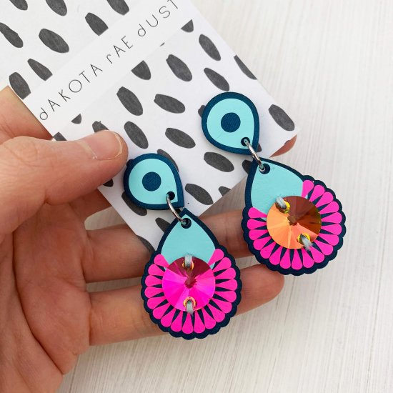 A pair of neon pink and peacock mini jewel earrings mounted on a black and white patterned, dakota rae dust branded card are held in a white woman's open palm