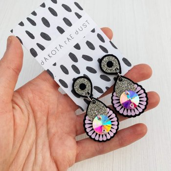A pair of black, lilac and silver glitter mini jewel earrings mounted on a black and white patterned, dakota rae dust branded card are held in a white woman's open palm