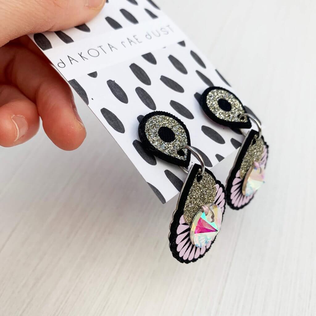 A pair of glittery mini jewel earrings mounted on a black and white patterned, dakota rae dust branded card held at a jaunty angle in a white hand