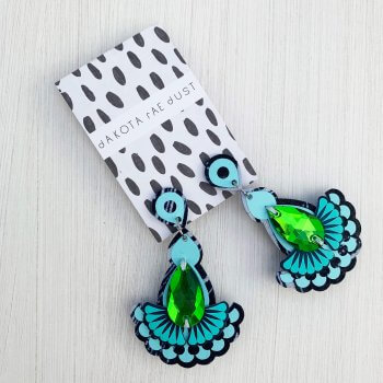 A pair of navy, pale blue, turquoise and green statement jewel earrings with iridescent teardrop shaped jewels, mounted on a black and white patterned, dakota rae dust branded card are seen against an off white textured background