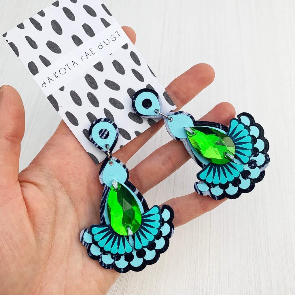 A pair of bright blue. green and turquoise Statement earrings with a shiny green jewel mounted on a black and white patterned dakota rae dust branded card are held in a woman's open hand