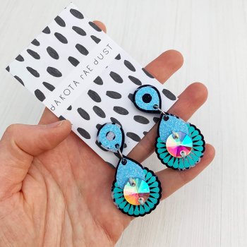 A pair of pale blue and turquoise mini jewel earrings mounted on a black and white patterned, dakota rae dust branded card are held in a white woman's open palm