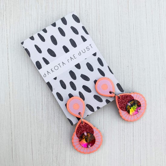 A pair of peach, lilac and pink glitter mini jewel earrings mounted on a black and white patterned, dakota rae dust branded card are seen against an off white plain background
