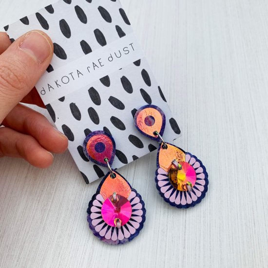 A pair of lilac and iridescent mini jewel earrings mounted on a black and white patterned, dakota rae dust branded card are held in a white woman's hand