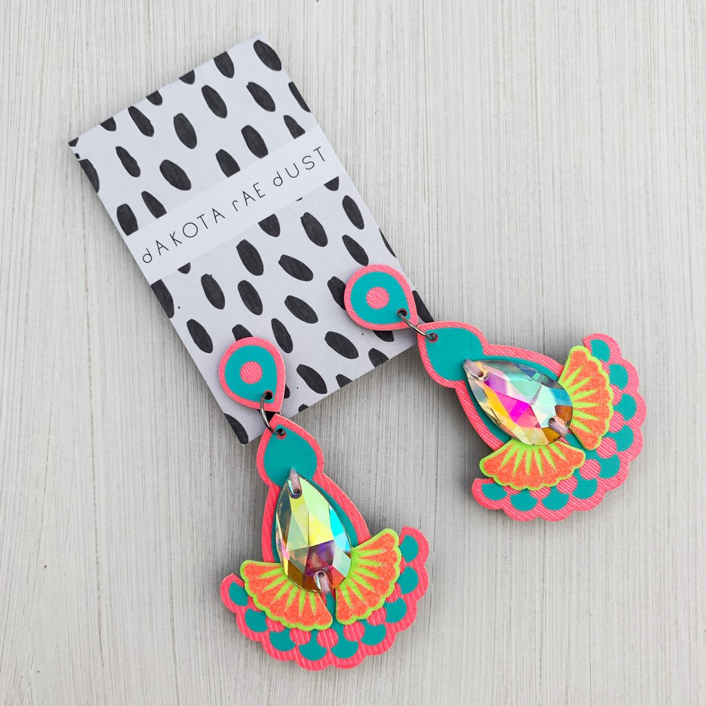 A pair of pink, turquoise, lime and orange statement jewel earrings with iridescent teardrop shaped jewels, mounted on a black and white patterned, dakota rae dust branded card are seen against an off white textured background