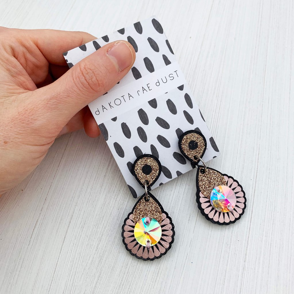 A pair of dark grey, metallic blush, gold glitter and iridescent mini jewel earrings mounted on a black and white patterned, dakota rae dust branded card are held in a white woman's hand
