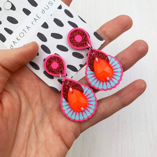 A pair of bright pink, glittery red and baby blue mini dangly earrings with teardrop shaped bright orange gems are held towards the camera in a woman's hand
