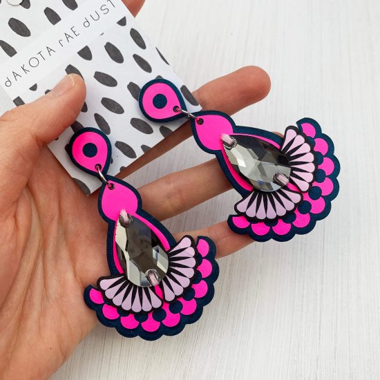 A pair of luxurious looking fluorescent pink statement earrings featuring teardrop shaped glass grey jewels are displayed on a black and white patterned, dakota rae dust branded card, held in a woman's hand