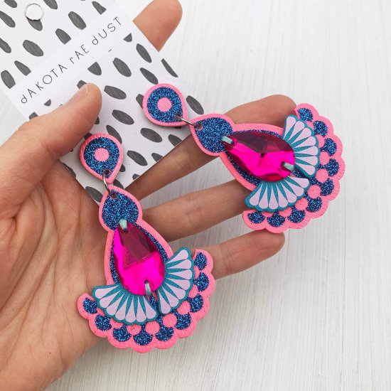 A pair of neon pink and glittery blue statement earrings featuring teardrop shaped glass hot pink jewels are displayed on a black and white patterned, dakota rae dust branded card, held in a woman's hand