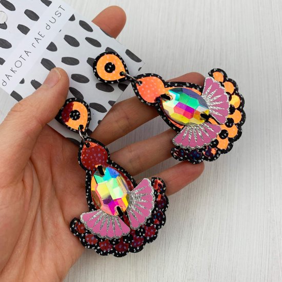 A pair of black, silver and pink iridescent colour change statement earrings featuring oval shaped glass iridescent jewels are displayed on a black and white patterned, dakota rae dust branded card, held in a woman's hand
