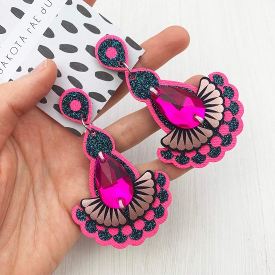 A pair of pink, peacock green glitter and blush statement earrings featuring teardrop shaped glass hot pink jewels are displayed on a black and white patterned, dakota rae dust branded card, held in a woman's hand