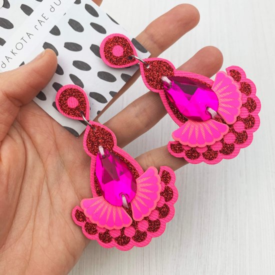 A pair of bright pink and red glitter statement earrings featuring teardrop shaped glass hot pink jewels are displayed on a black and white patterned, dakota rae dust branded card, held in a woman's hand