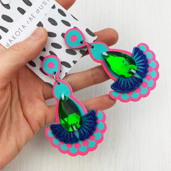 A pair of pink, turquoise and blue statement earrings featuring teardrop shaped glass emerald green jewels are displayed on a black and white patterned, dakota rae dust branded card, held in a woman's hand