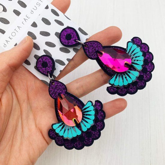 A pair of purple glitter, navy and turquoise statement earrings featuring teardrop shaped glass dark iridescent jewels are displayed on a black and white patterned, dakota rae dust branded card, held in a woman's hand