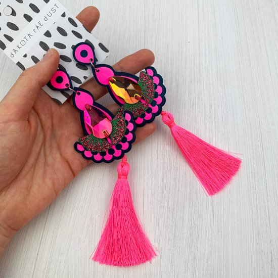 A pair of fluorescent pink and teal statement tassel earrings adorned with dark iridescent colour change jewels are seen laid out on the palm of a white woman's open hand