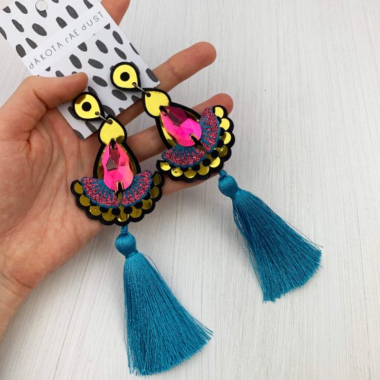 A pair of bright blue and gold statement tassel earrings adorned with dark iridescent colour change jewels are seen laid out on the palm of a white woman's open hand