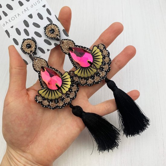 A pair of black and warm glittery gold statement tassel earrings adorned with dark iridescent colour change jewels are seen laid out on the palm of a white woman's open hand