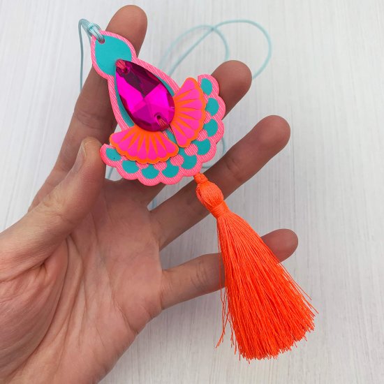 a fluorescent pink and turquoise statement tassel pendant necklace with a hot pink teardrop shaped jewel is held towards the camera in a white woman's hand