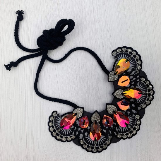 A black and silver statement jewel necklace with an ornate bib decorated with dark iridescent jewels. The necklace is lying flat on an off white background