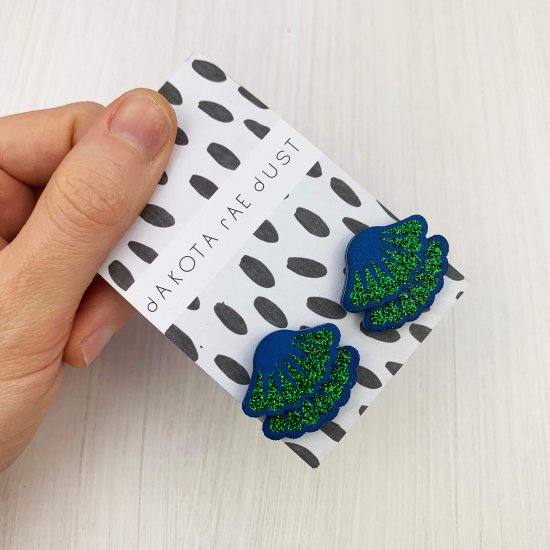 A pair of blue and glittery green tiered frill stud earrings mounted on a black and white patterned card are held in a woman's hand