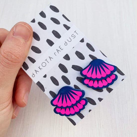 A pair of royal blue and neon pink Tiered frill stud earrings are mounted on a black and white patterned, dakota rae dust branded card and held in a woman's thumb and fore finger