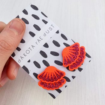 A pair of bright red and glittery pink Tiered frill stud earrings are mounted on a black and white patterned, dakota rae dust branded card and held in a woman's thumb and fore finger