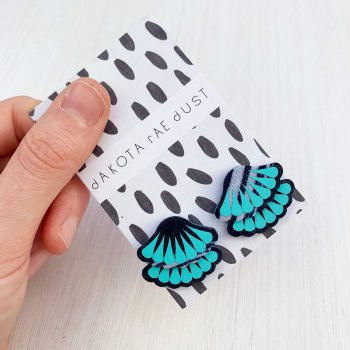 A pair of navy, white and turquoise Tiered frill stud earrings are mounted on a black and white patterned, dakota rae dust branded card and held in a woman's thumb and fore finger