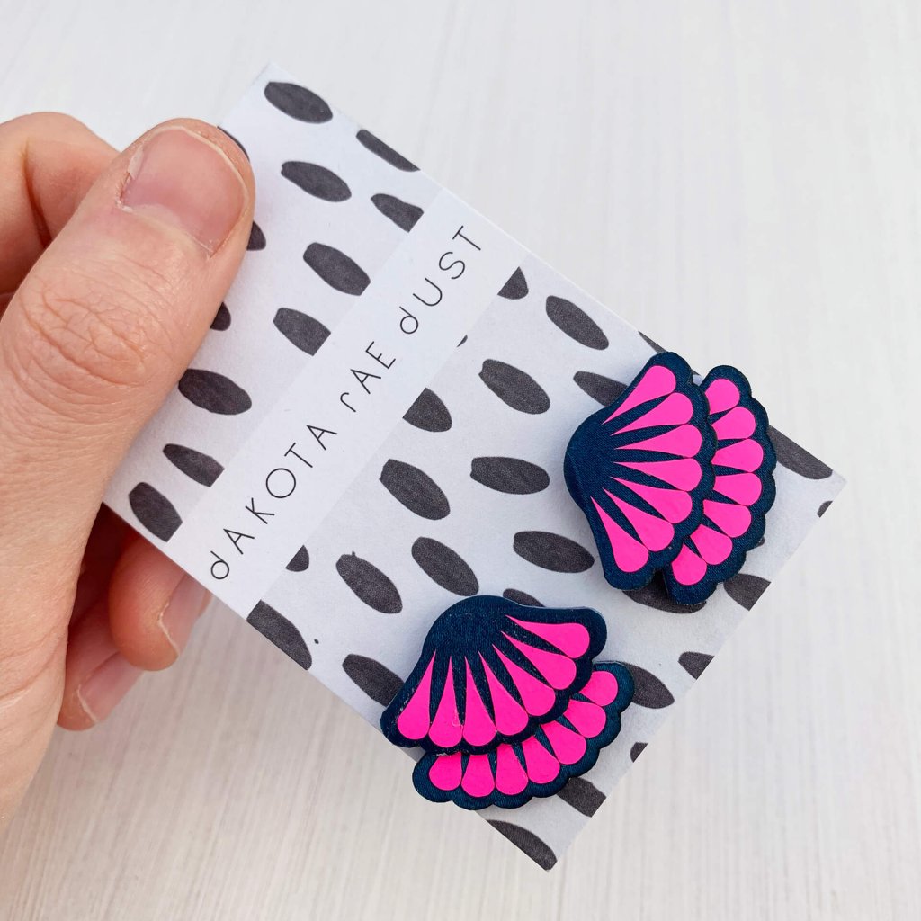 A pair of peacock green and neon pink Tiered frill stud earrings are mounted on a black and white patterned, dakota rae dust branded card and held in a woman's thumb and fore finger