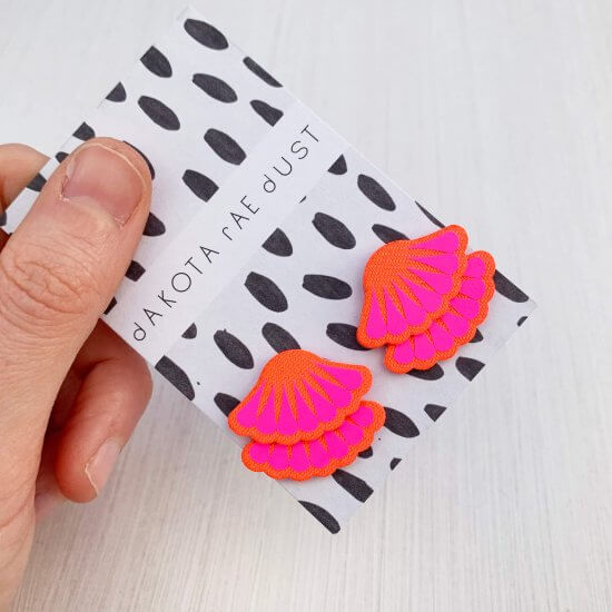 A pair of bright red and neon pink Tiered frill stud earrings are mounted on a black and white patterned, dakota rae dust branded card and held in a woman's thumb and fore finger