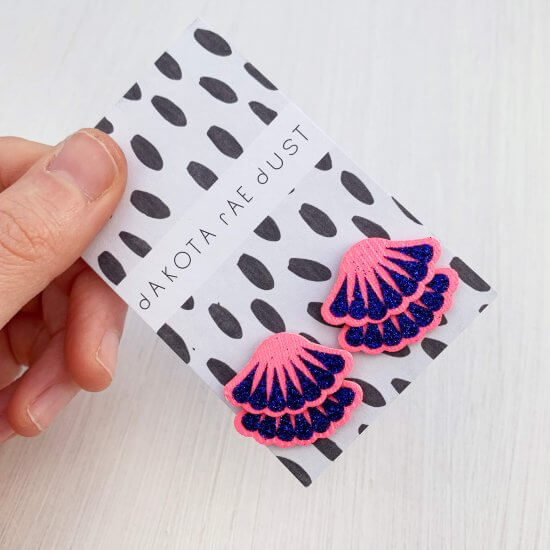 A pair of neon pink and glittery royal blue Tiered frill stud earrings are mounted on a black and white patterned, dakota rae dust branded card and held in a woman's thumb and fore finger