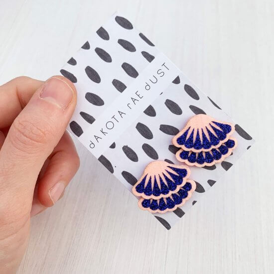 A pair of pale pink and glittery royal blue tiered frill stud earrings mounted on a black and white patterned, dakota rae dust branded card, held between a white thumb and fore finger.