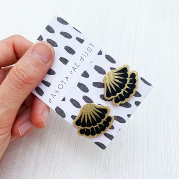 a pair of tiered stud earrings in black and gold, mounted on a black and white patterned, dakota rae dust branded card and held against an off white background between a white thumb and forefinger