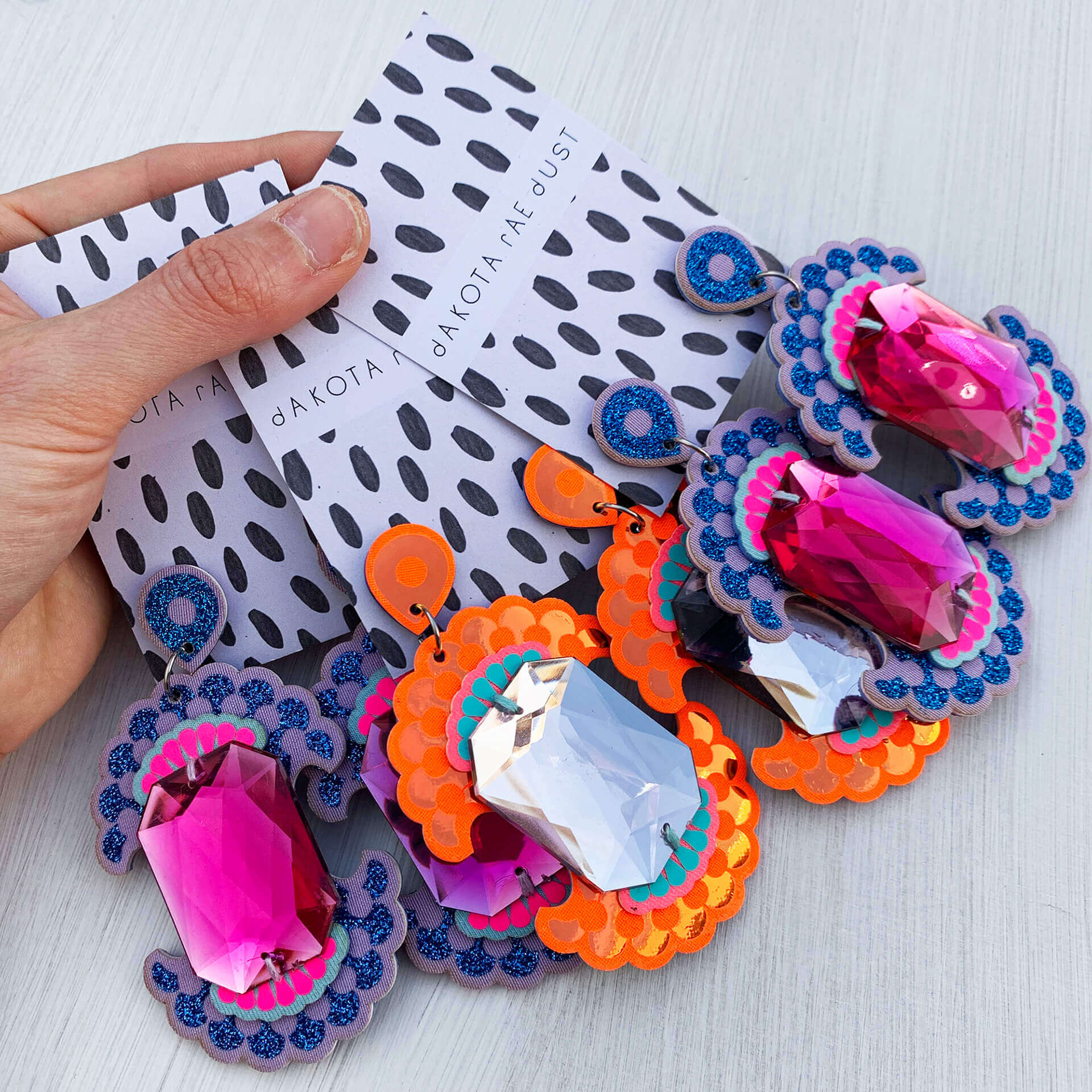 Three pairs over colourful, oversize earrings adorned with giant jewels mounted on black and white patterned, dakota rae dust branded cards are held in a woman's hand