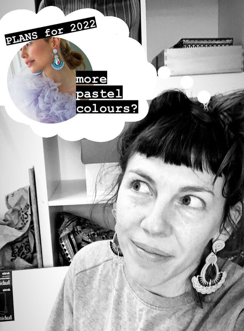 A black and white photo of dakota rae dust designer Bec, a white woman with a short fringe, looking off to one side of the photo with a white cartoon dream style cloud above her head. The cloud contains a photo of some blue and lilac earrings modelled by a young woman and the words "PLANS for 2022, more pastel colours?"