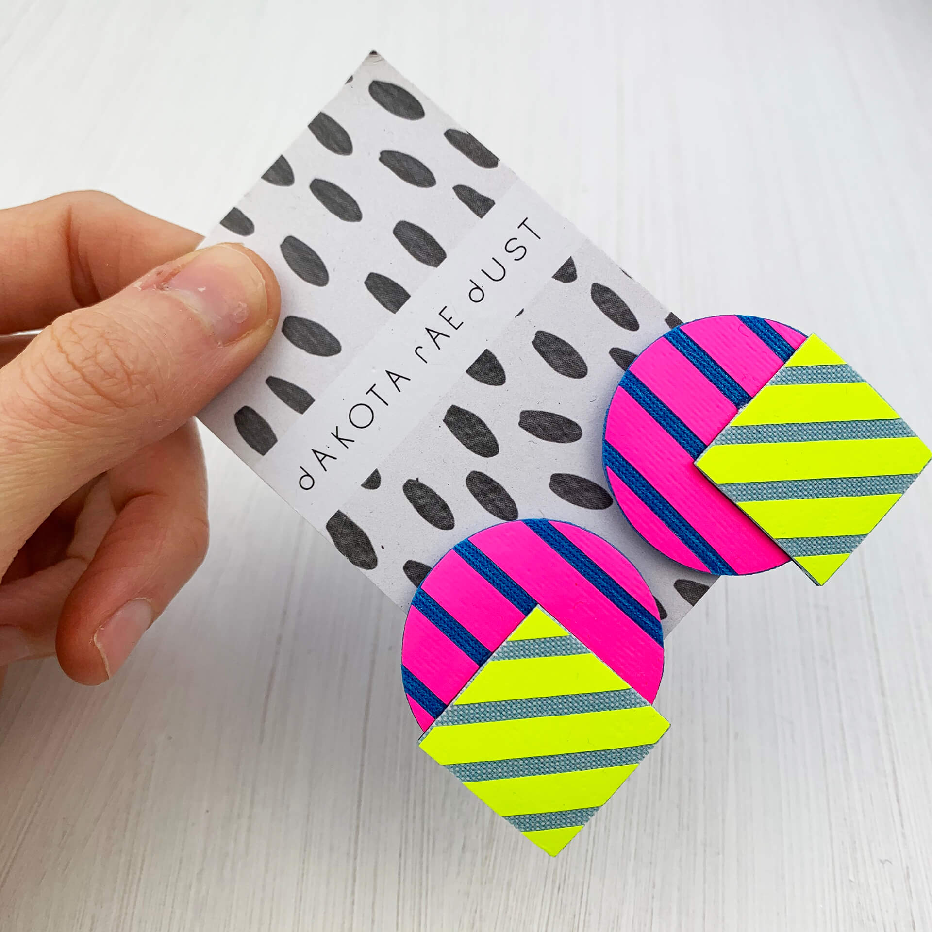 A pair of large colourful stud earrings made of a circle and square printed with fluorescent graphic stripes. The earrings are mounted on a black and white patterned, dakota rae dust branded card which is held between a woman's thumb and forefinger against an off white background
