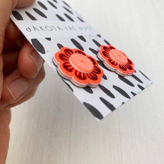 a pair of orange, red and peach flower shaped studs, mounted on a black and white patterned, dakota rae dust branded card and held by some just visible fingers