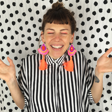 A white woman wearing a black and white striped shirt and large fluorescent orange tassel earrings is facing the camera smiling. Her eyes are closed and her hands are raised up, palms open. The background of the image is a black and white polka dot pattern.