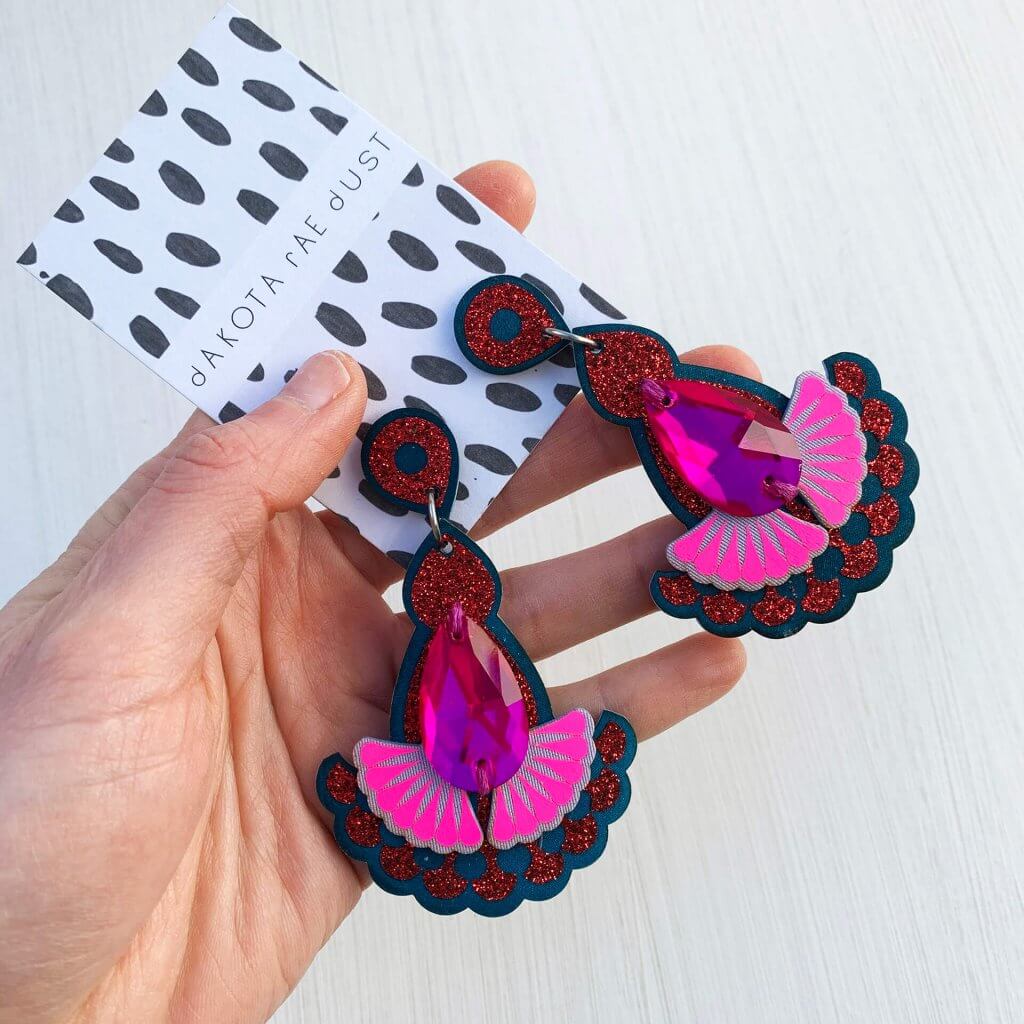 A pair of jewelled statement earrings in teal, red glitter, lilac and fluorescent pink mounted on a black and white patterned dakota rae dust card are held in a white woman's hand against an off white background.