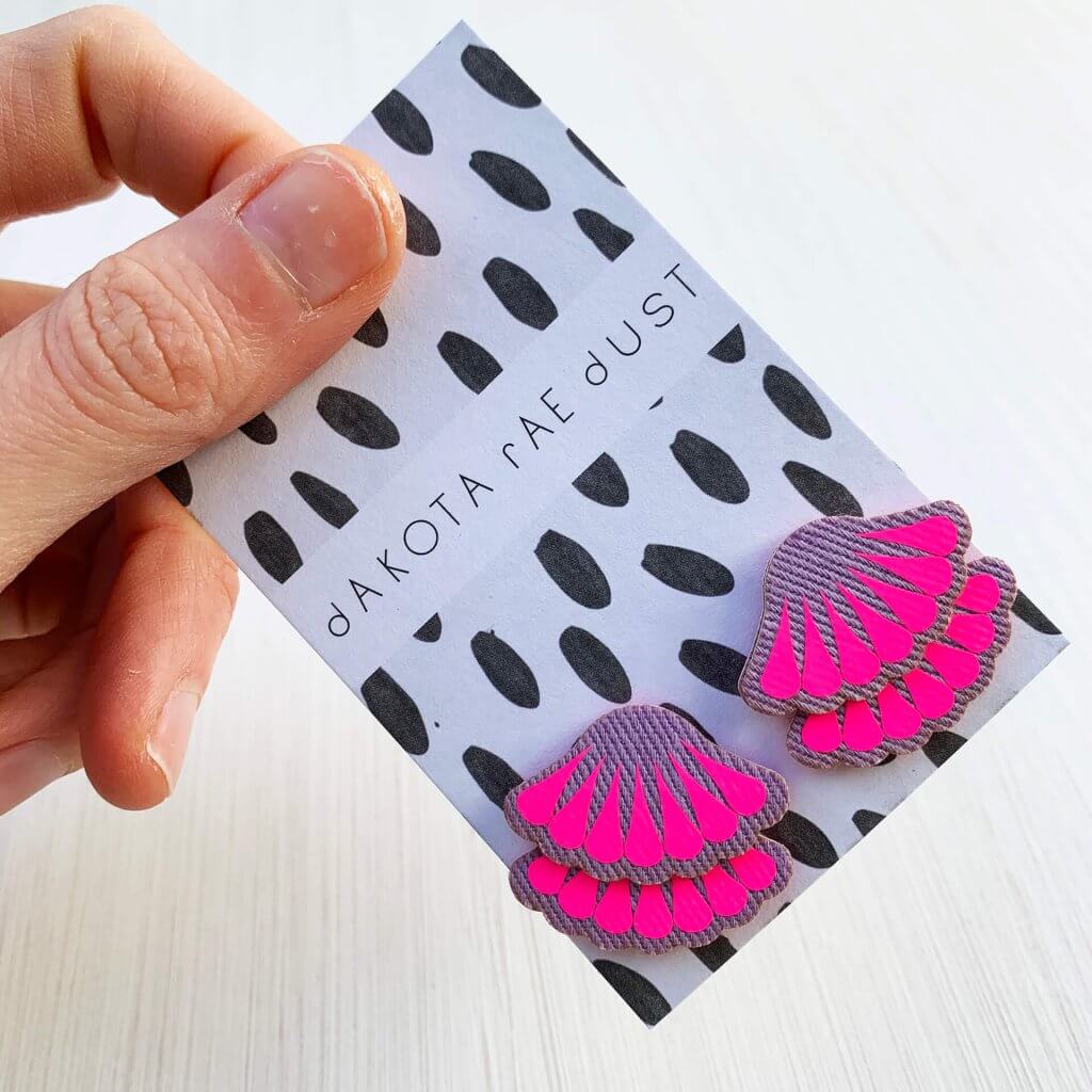 A pair of lilac and fluorescent pink frill stud earrings mounted on a black and white patterned, dakota rae dust branded card are held between the thumb and forefinger of a woman's hand against an off white background