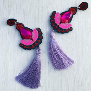 A pair of lilac, teal, red glitter and fluorescent pink Tassel statement earrings with teardrop shaped pink jewels are lying on an off white background