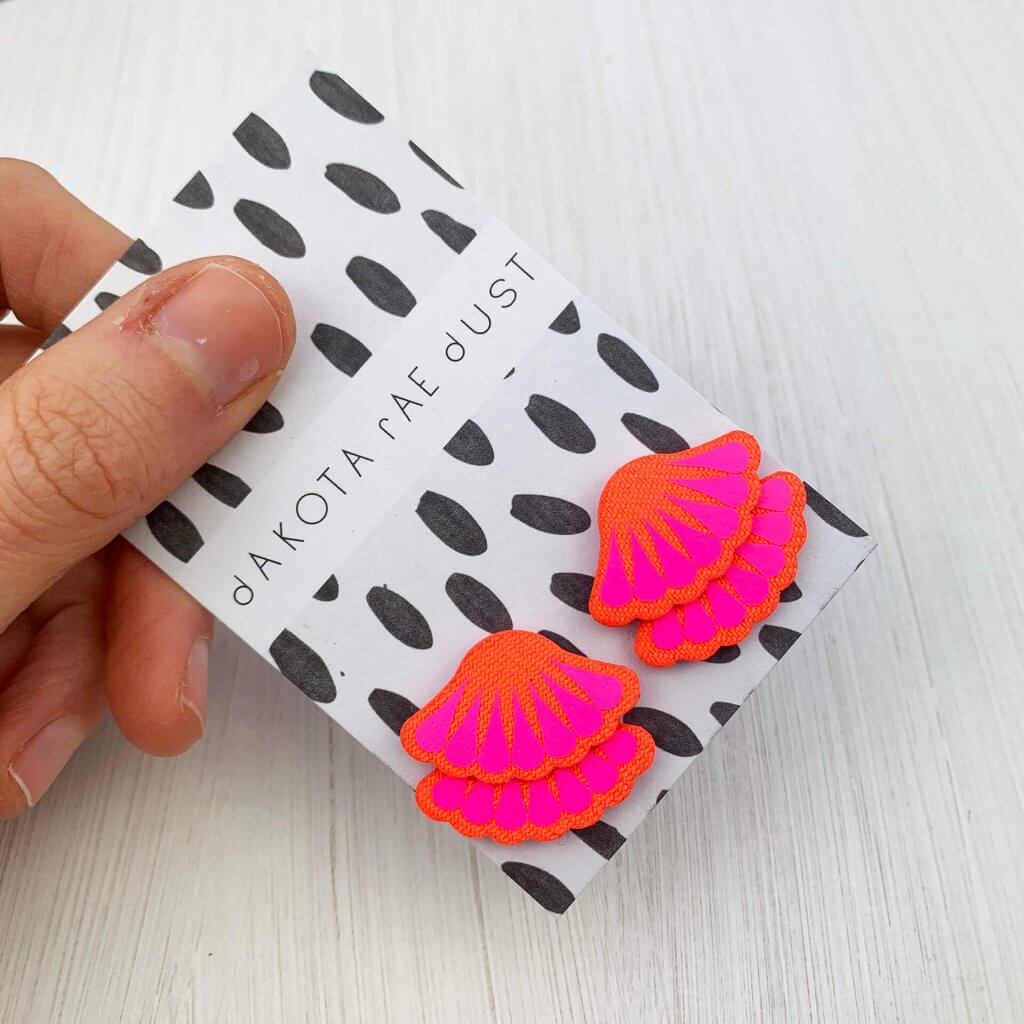A pair of hot pink frill stud earrings backed on a black and white patterned dakota rae dust branded card, held in a woman's thumb and forefinger against a white background