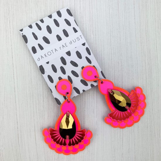 A pair of hot pink earrings with a gold jewel are mounted on a black and white patterned, dakota rae dust branded card are seen lying on an off white textured background