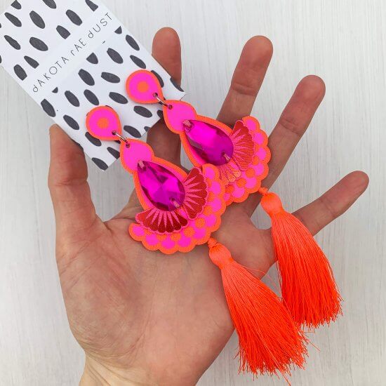 A pair of hot pink and orange statement tassel earrings are held in an open hand