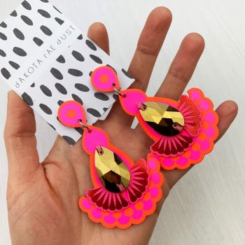 A pair of bright pink, red and gold statement earrings held in a woman's hand