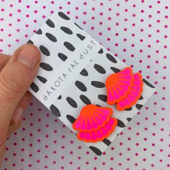 A pair of hot pink frill stud earrings backed on a black and white patterned dakota rae dust branded card, held in a woman's thumb and forefinger against a white background with tiny pink polka dots
