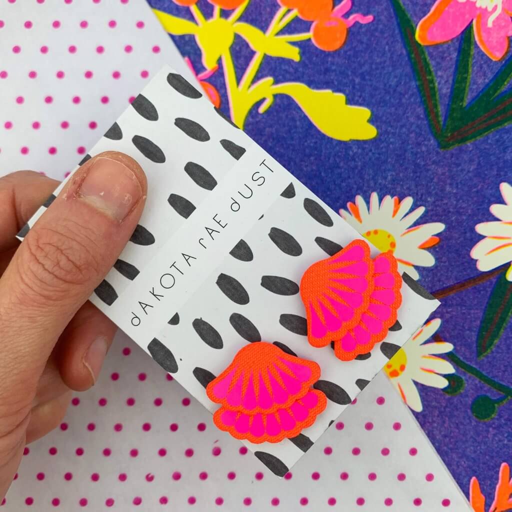 A pair of hot pink frill stud earrings mounted on a black and white patterned, dakota rae dust branded card are held between the thumb and forefinger of a woman's hand against a colourful background.