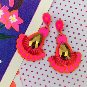A pair of hot pink earrings with a gold jewel are seen against a colourful background consisting of a royal blue floral patten and an area of pink and white polka dot pattern